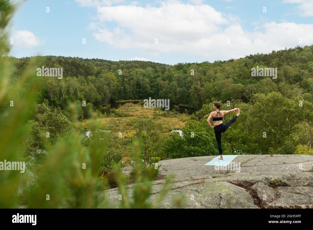 Nicht erkennbares Weibchen mit der Hand auf der Taille tut Extended Hand zu Big Toe Pose auf Stein in üppiger Landschaft im Sommer Stockfoto