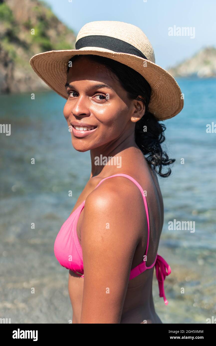 Seitenansicht einer positiven Frau im Sommer-Outfit und mit aufblasbarer Matratze, die an sonnigen Tagen im Urlaub an der sandigen Küste spazieren geht Stockfoto