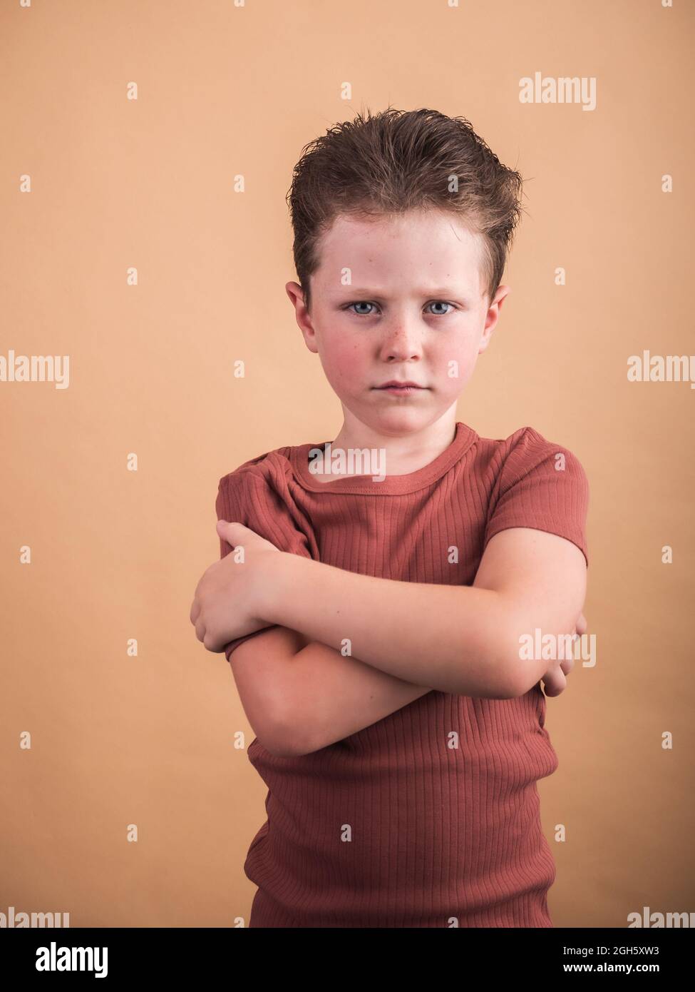 Unzufriedenes Kind mit gefalteten Armen und braunem Haar, das auf beigem Hintergrund auf die Kamera blickt Stockfoto