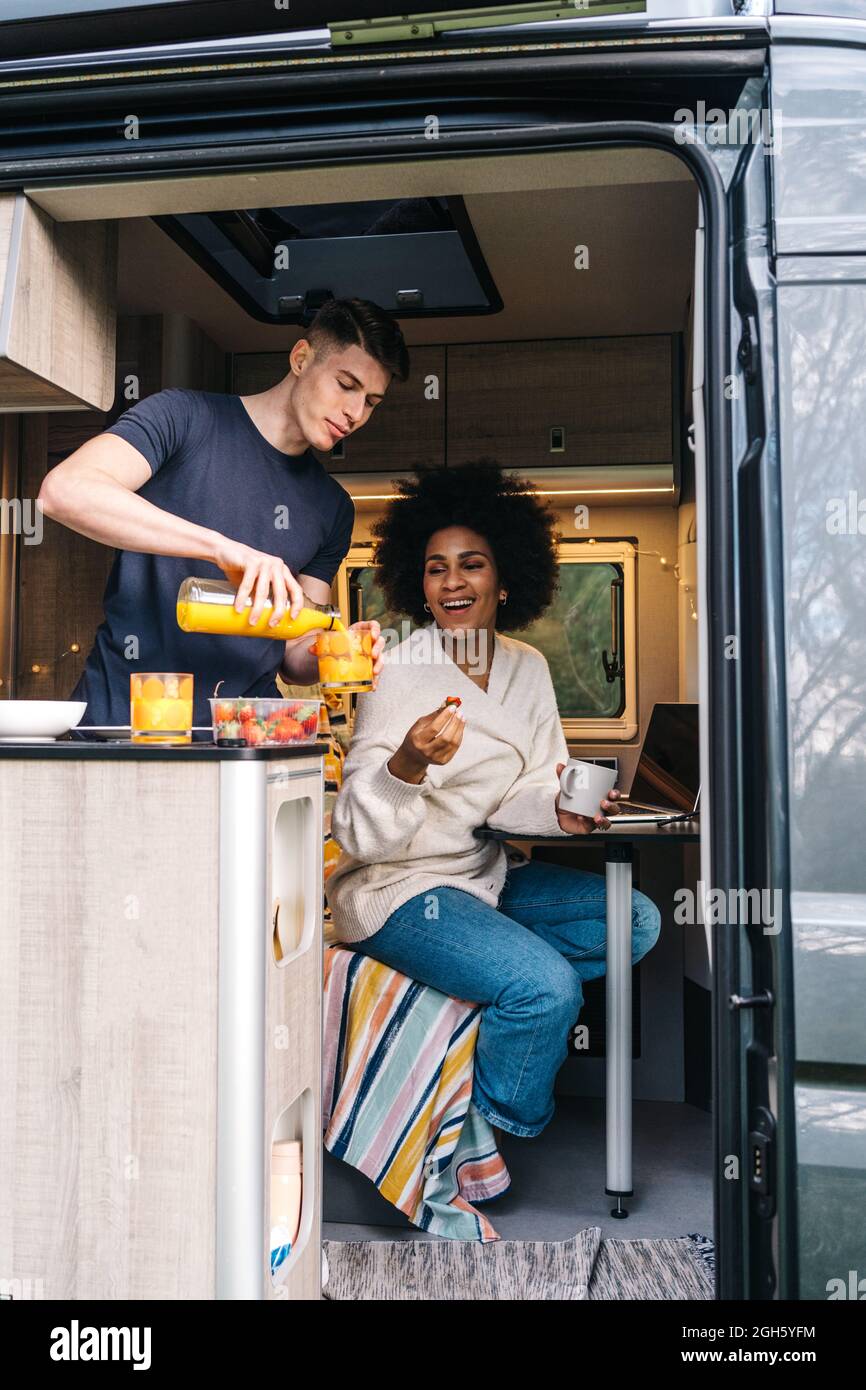 Junger Mann serviert natürlichen Saft zu fröhlichen schwarzen Freundin, während das Frühstück zusammen in Wohnmobil Anhänger während der Sommerferien genießen Stockfoto