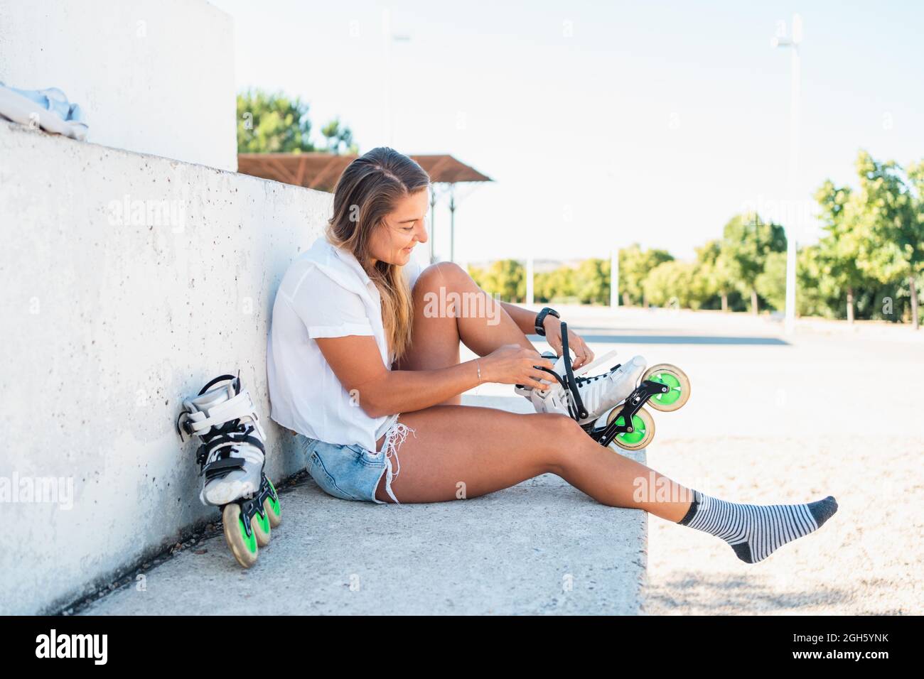 Seitenansicht einer fröhlichen Skaterin, die im Stadtgebiet auf dem Boden sitzt und Rollerblades aufsetzt, während sie das Wochenende im Sommer genießt Stockfoto