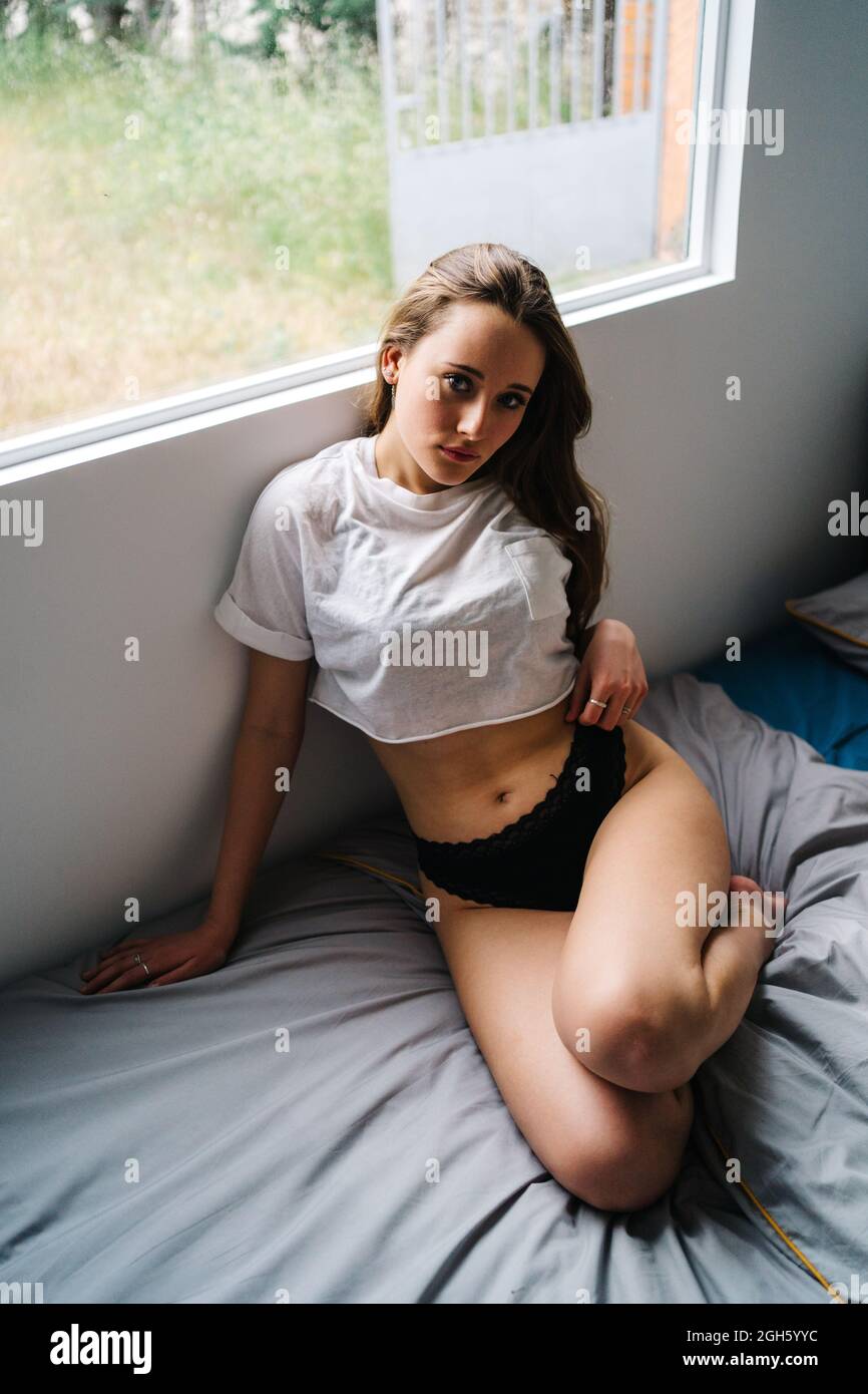 Seitenansicht einer zarten Frau, die den Hals berührt, während sie zu Hause auf einem weichen Bett in der Nähe des Fensters sitzt und die Kamera anschaut Stockfoto
