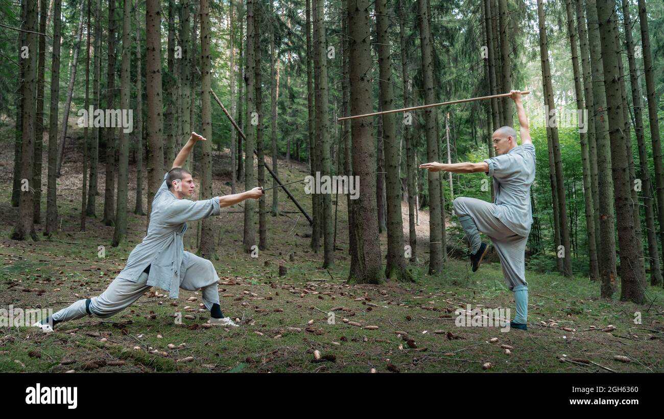 Ganzkörpermänner in grauen Kleidern üben beim Training im Wald Kung Fu mit Stock und Schwert Stockfoto