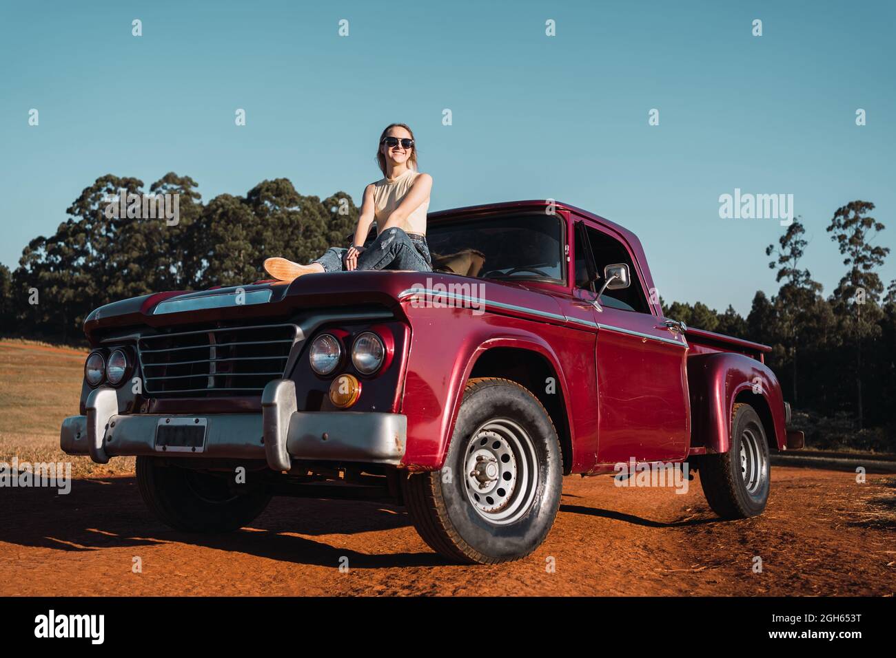 Niedriger Winkel der entzückten kühlen Frau sitzt auf der Haube der roten vintage Pickup Auto in der Landschaft im Sommerabend geparkt Stockfoto