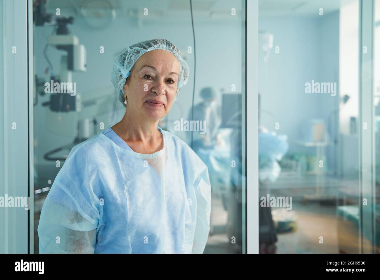 Freundliche Ärztin mittleren Alters in OP-Uniform, die die Kamera gegen einen nicht erkennbaren Kollegen im Krankenhaus anschaut Stockfoto