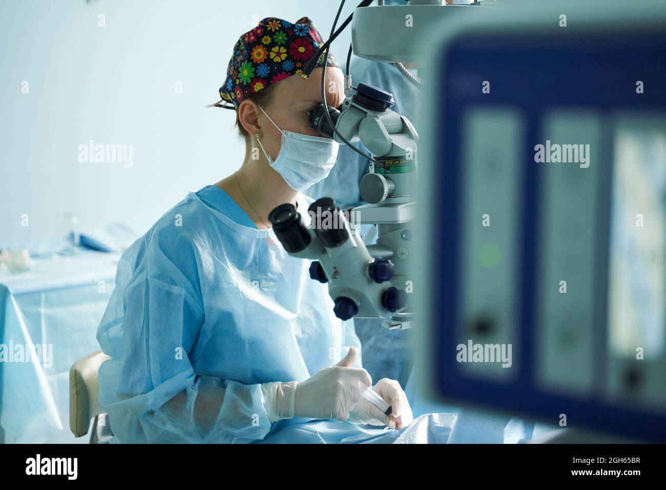 Aufmerksame Ärztin in einer chirurgischen Uniform und sterilen Maske, die durch das Mikroskop schaut, während sie das Auge eines nicht erkennbaren Patienten im Krankenhaus operiert Stockfoto