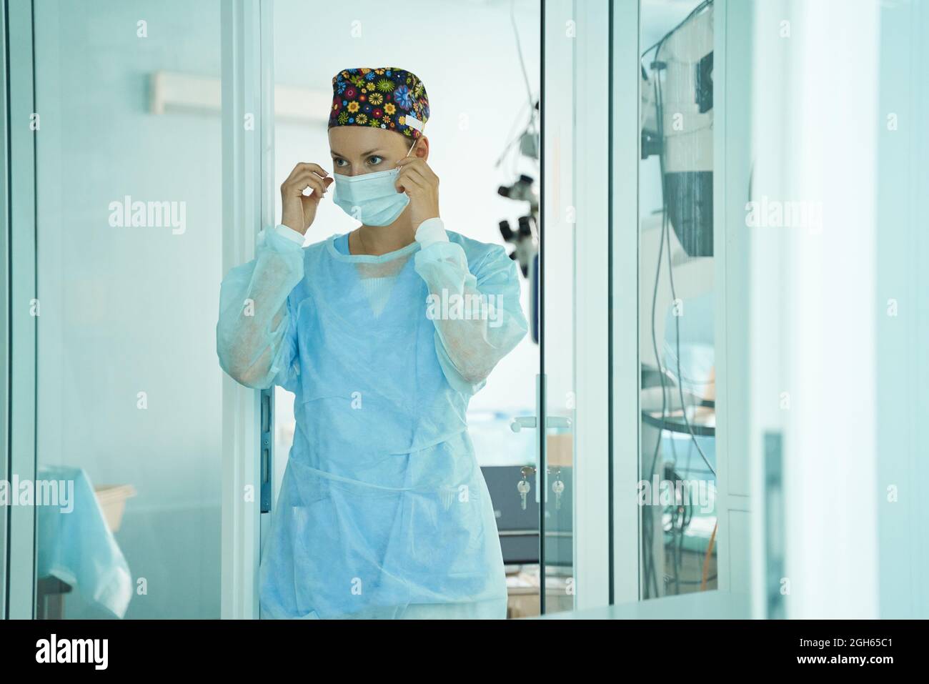 Erwachsene Ärztin in chirurgischer Uniform und ornamentaler medizinischer Kappe, die Einwegmaske aufsetzt, während sie im Krankenhaus nach vorne blickt Stockfoto