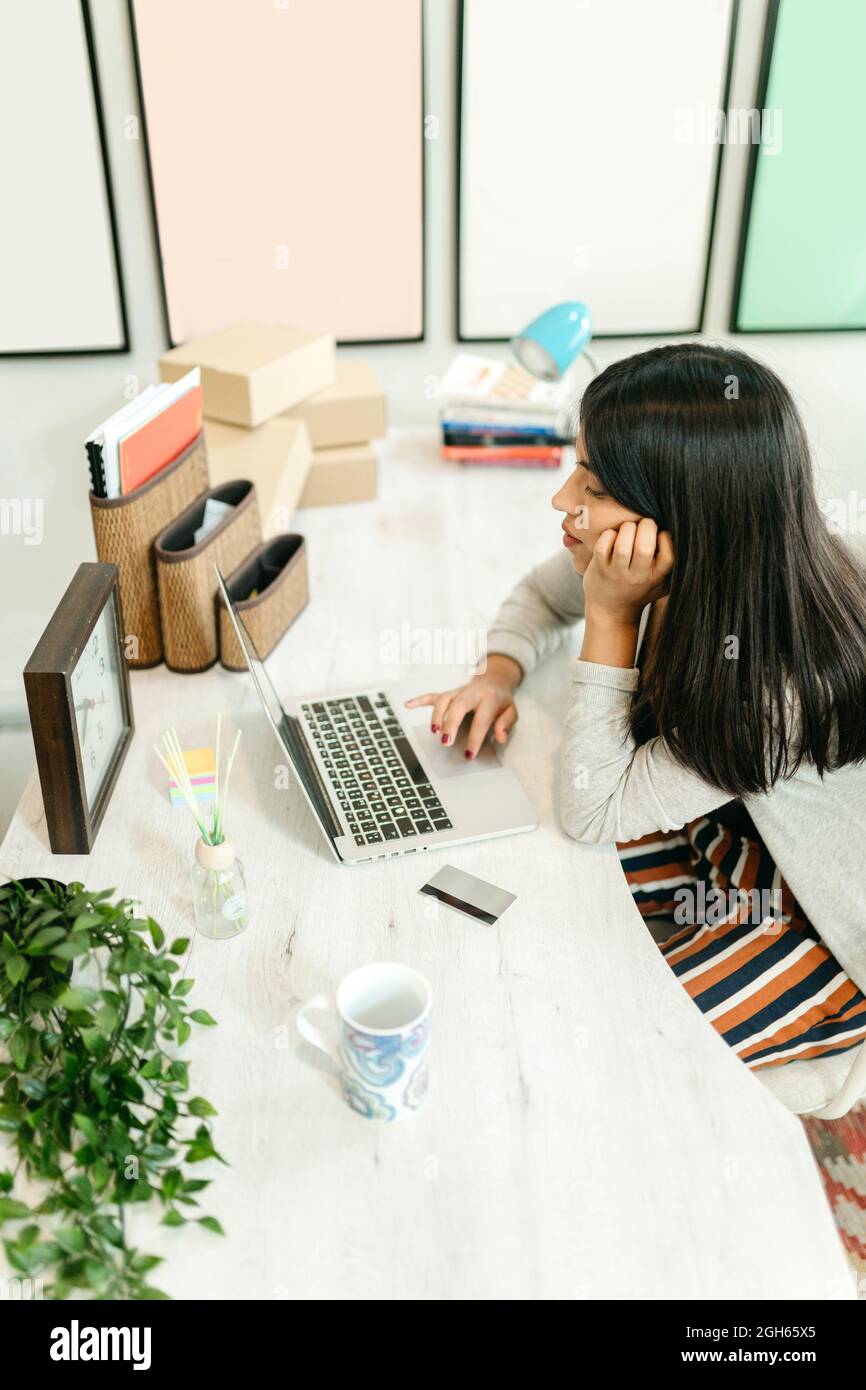 Seitenansicht von oben auf die Frau, die beim Online-Shopping Netbook benutzt, während sie zu Hause am Tisch mit Plastikkarte sitzt und Waren auswählt Stockfoto