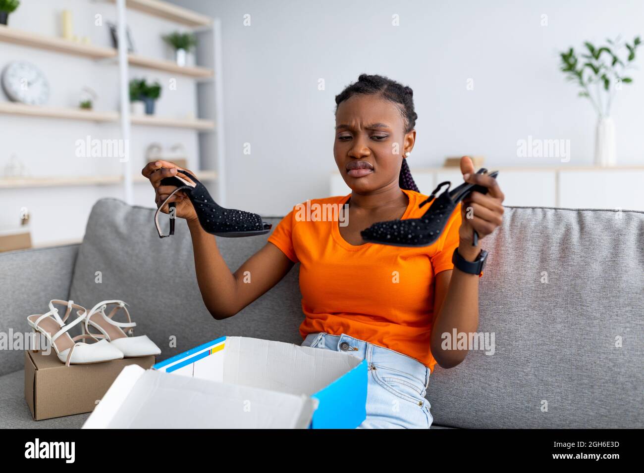 Enttäuschte Afro-Dame beim Auspacken des Kartonpakets, beim Empfang von Schuhen, unglücklich über das gelieferte Produkt, vor Ekel knirschende Nase Stockfoto