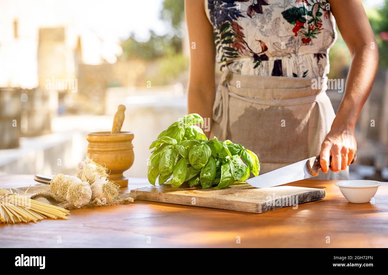 Hände einer Frau hacken grünes Basilikum am Küchentisch in einem Garten im Freien Stockfoto