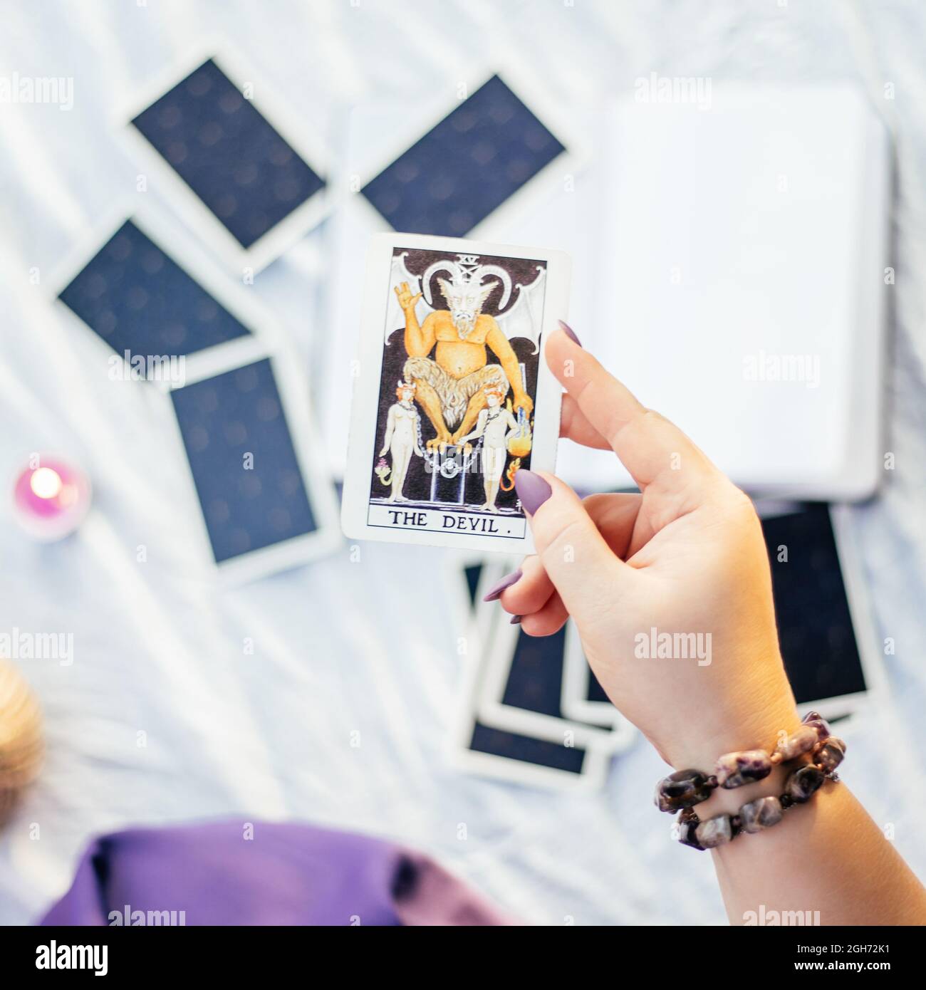 Weibliche Hand mit lila Nägeln hält Tarot-Karte namens der Teufel über der weißen Oberfläche mit offenem Notizbuch und Kerze. Draufsicht. Minsk, Weißrussland - 07.28.2021 Stockfoto