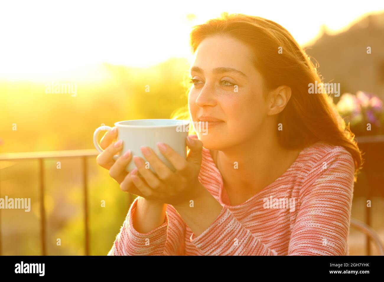 Eine zufriedene Frau, die beim Sonnenuntergang auf einem Balkon die Aussicht mit einem Kaffeebecher betrachtet Stockfoto