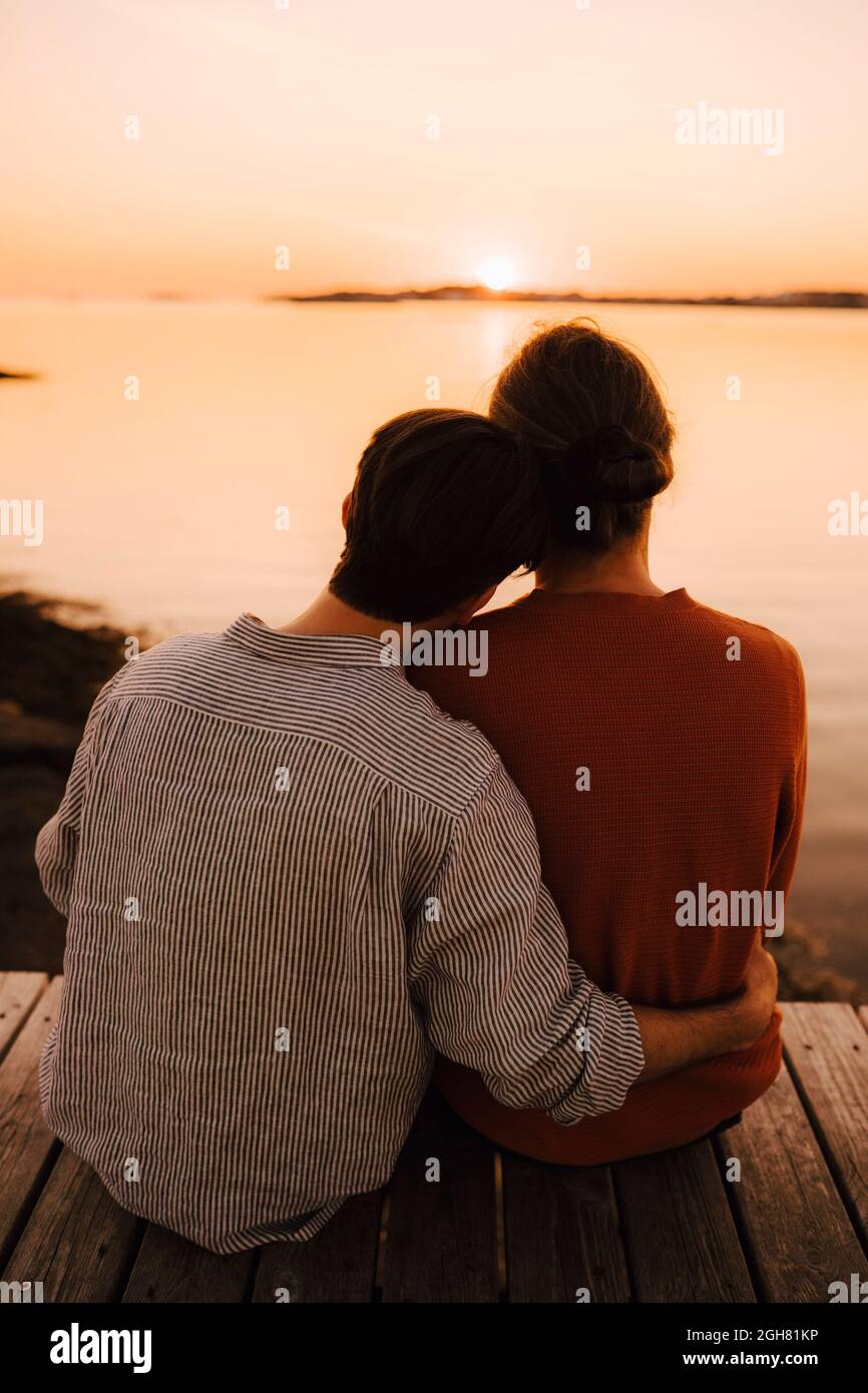 Rückansicht von männlichen Freunden, die den Sonnenuntergang betrachten, während sie auf dem Pier sitzen Stockfoto