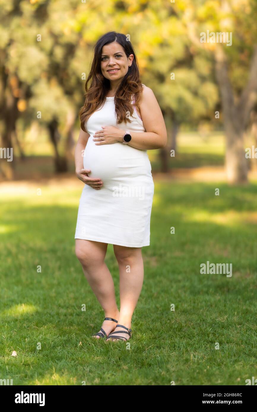 Vertikales Foto einer lächelnden Schwangeren, die im Freien steht und ihren Bauch berührt Stockfoto