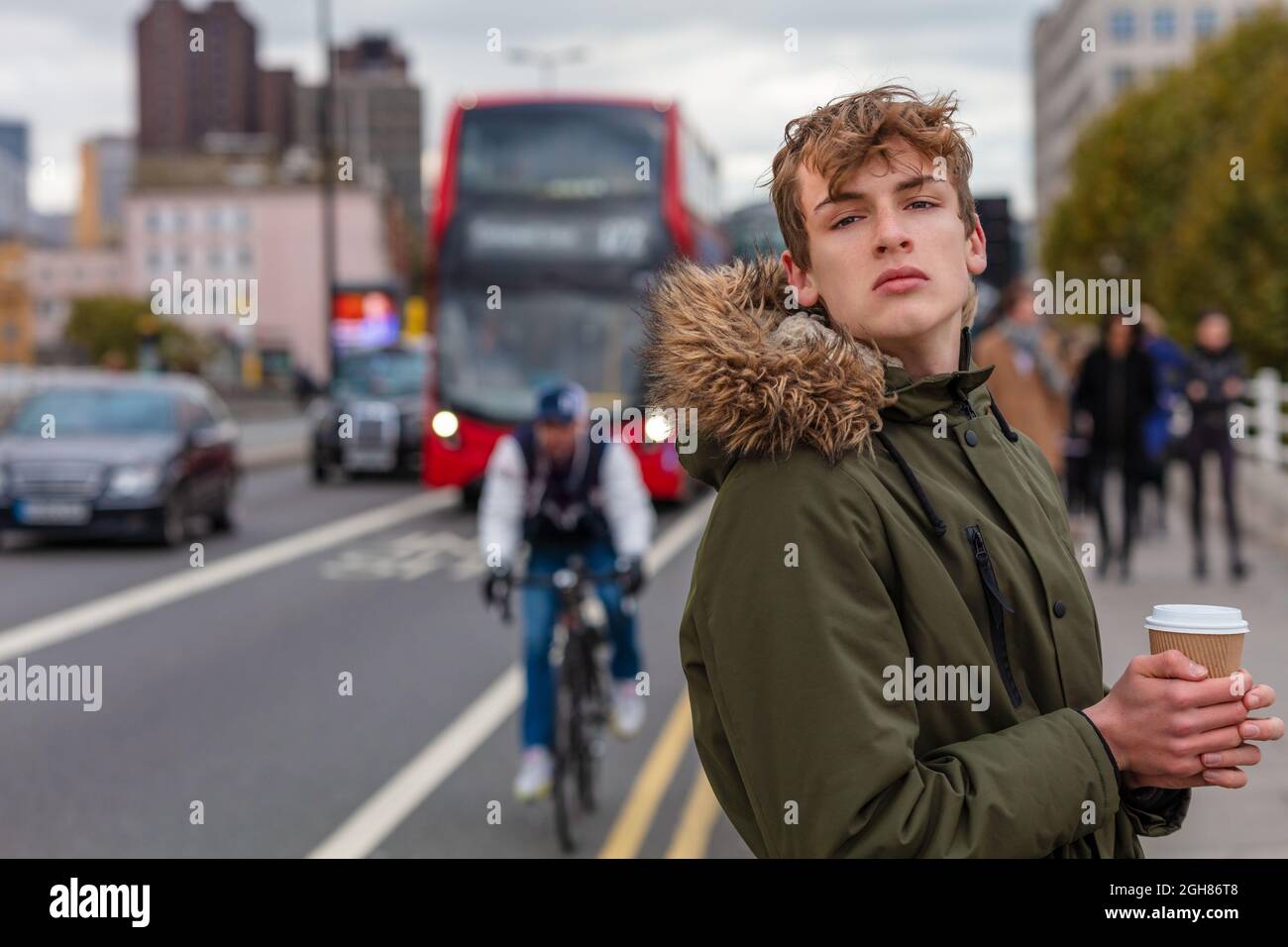 Junger Teenager mit Parka-Jacke vor dem Hotel, der Kaffee von einem roten Londoner Bus, Autos, einem Radfahrer, Fußgängern und dem Verkehr trinkt Stockfoto