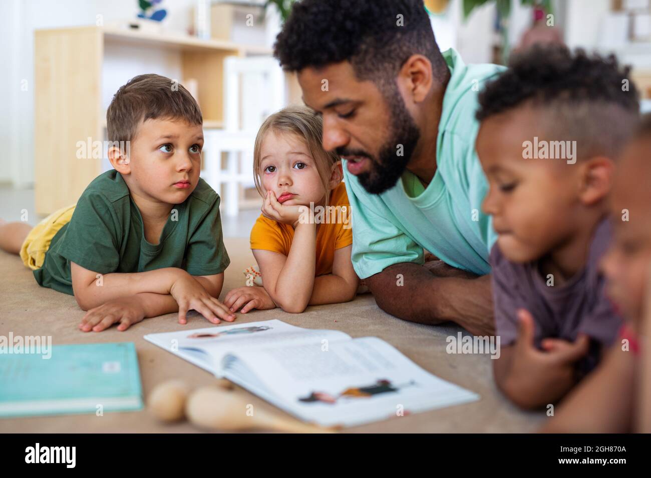 Gruppe von kleinen Kindergarten-Kinder mit Mann Lehrer auf dem Boden drinnen im Klassenzimmer, Buch zu lesen. Stockfoto