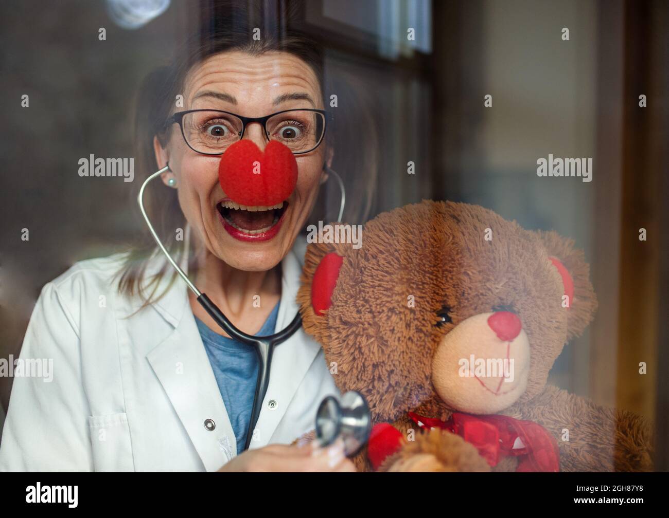 Porträt einer lächelnden Ärztin mit roter Clownnase, die einen Teddybären hält und die Kamera anschaut. Stockfoto