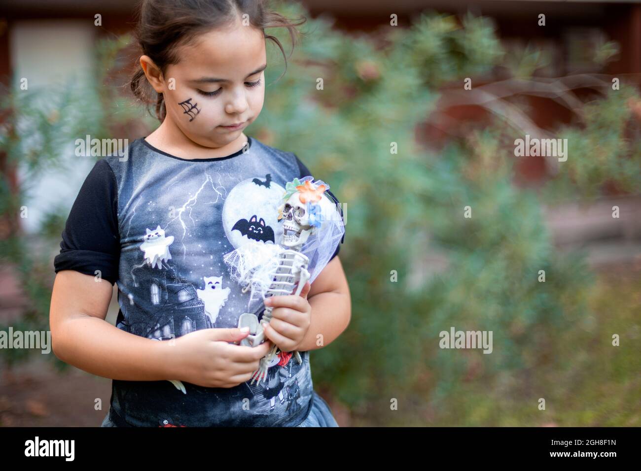 Kleines kaukasisches Kind, verkleidet als Hexe mit Spielzeugskelett in ihren Armen. Sie ist draußen. Platz für Text. Stockfoto