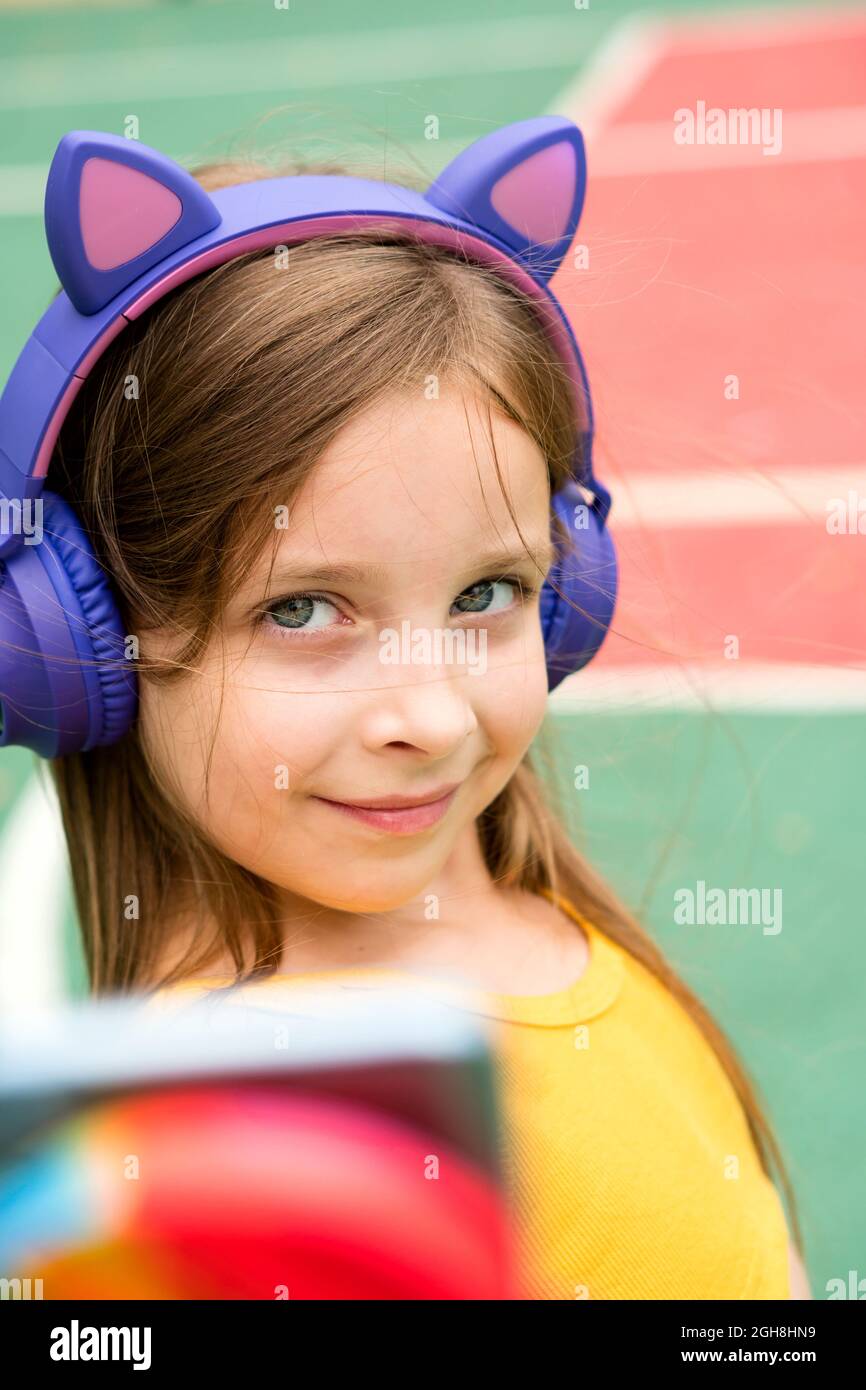 Sorglos froh Mädchen in kabellosen Kopfhörern nimmt Selfie streckt Arm nach vorne mit unkenntlichen Gerät geht im Freien chillen auf Sportplatz. Stockfoto