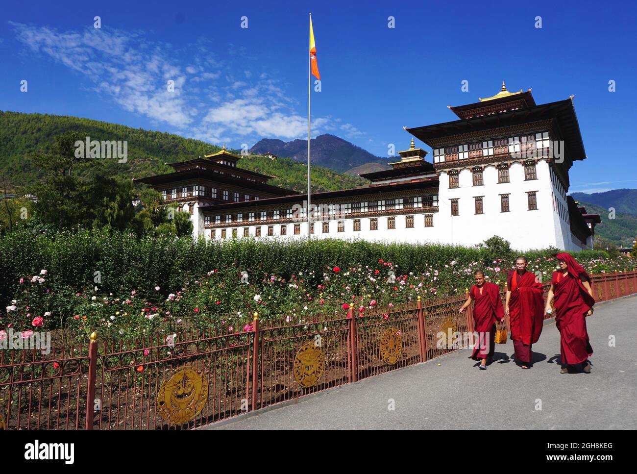 Drei buddhistische Mönche mit roten Roben gehen an wunderschönen Blumengärten vor dem Tashichho Dzong vorbei, dem offiziellen Sitz der Regierung Bhutans seit 1952. Stockfoto