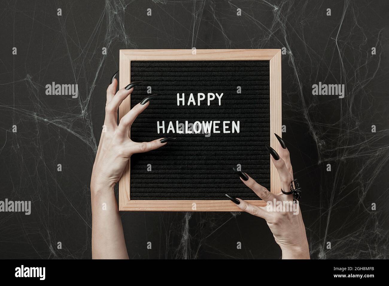 Happy Halloween auf Briefbrett in Händen mit langen schwarzen Nägeln und Spinnenring auf schwarzem Hintergrund mit Spinnweben. Halloween Feiertagskonzept. Stockfoto