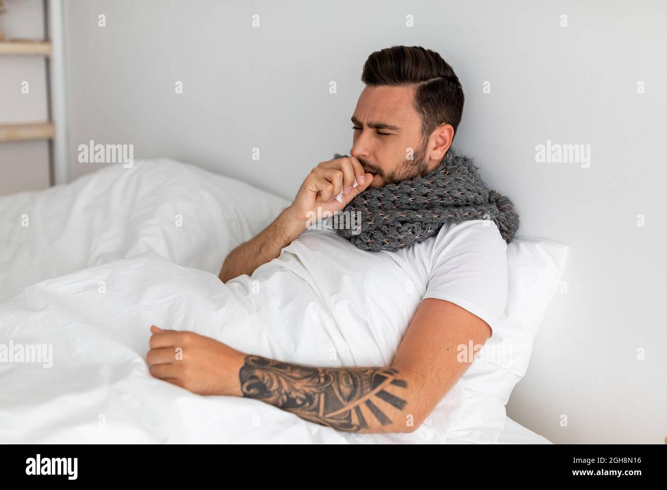 Covid-19. Kranker Kerl hustet, leidet an einer Lungenentzündung des Coronavirus, sitzt im Bett unter der Decke, freier Platz Stockfoto