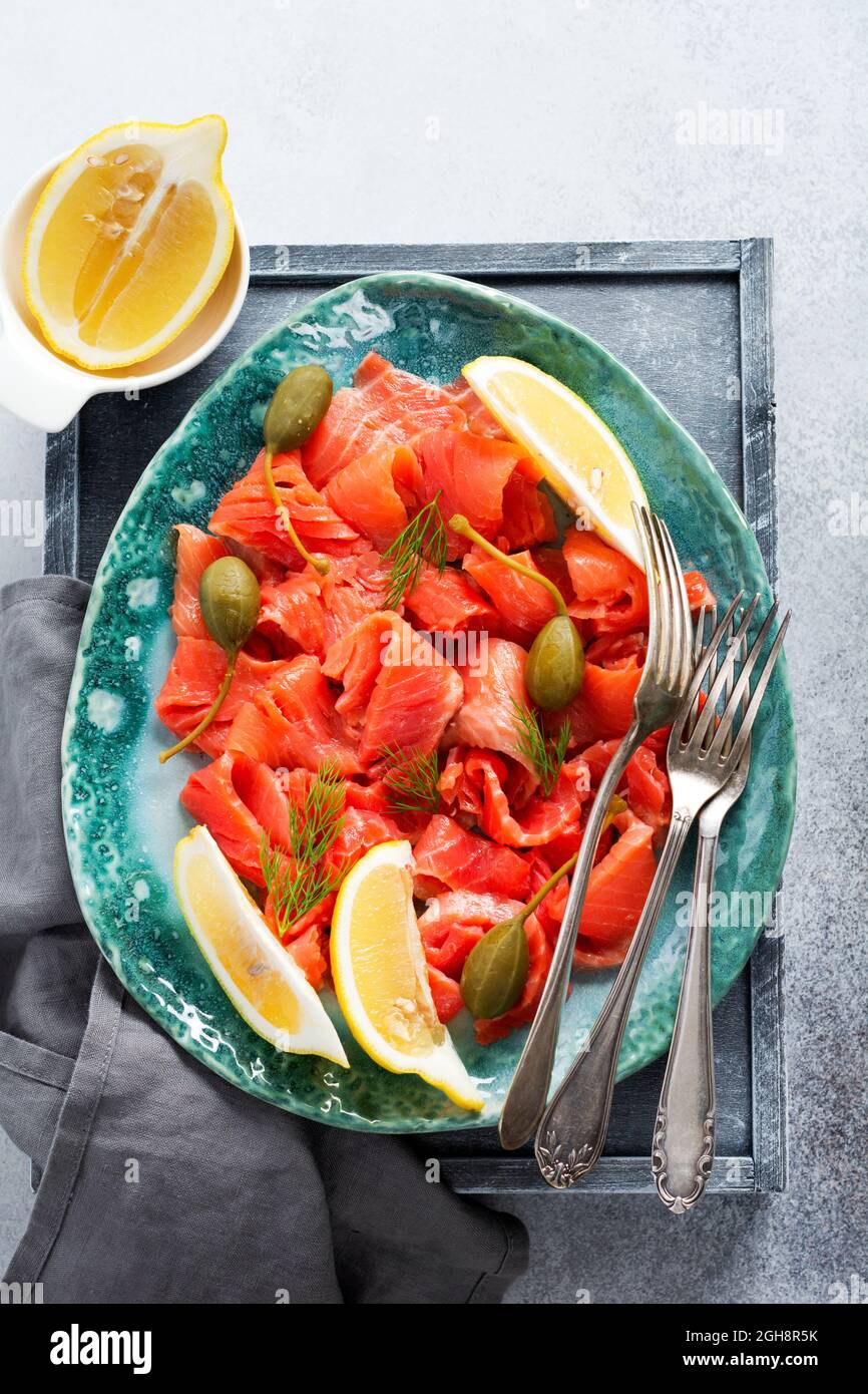 Scheiben leicht gesalzener Lachs mit Kapern, Zitrone und Dill auf einer Keramikplatte auf grauem Betonboden. Draufsicht Stockfoto