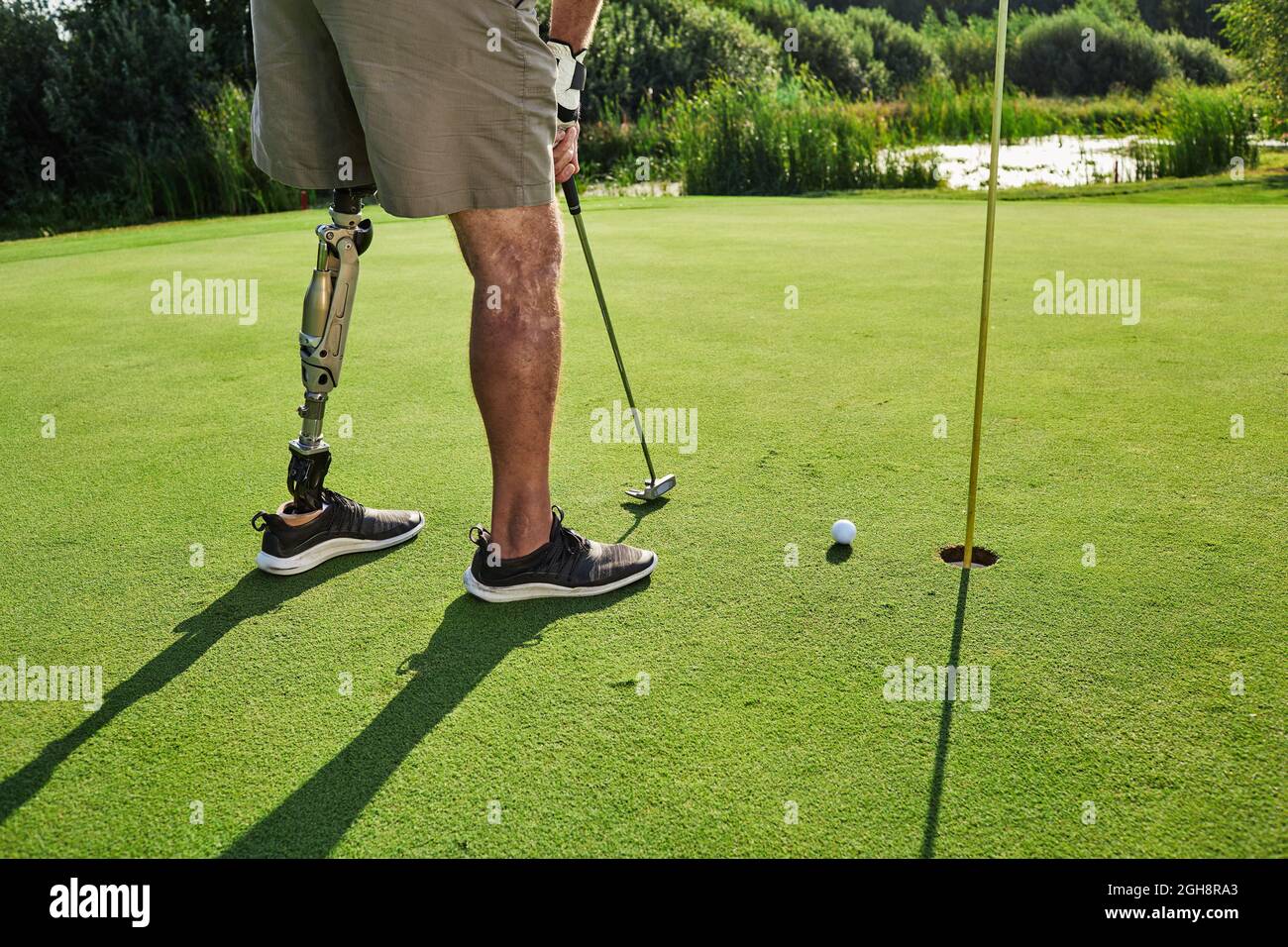 Professioneller Golfspieler mit Beinprothese trifft mit Putter auf den Golfball während des Golfspiels an sonnigen Tagen mit langen Schatten Stockfoto