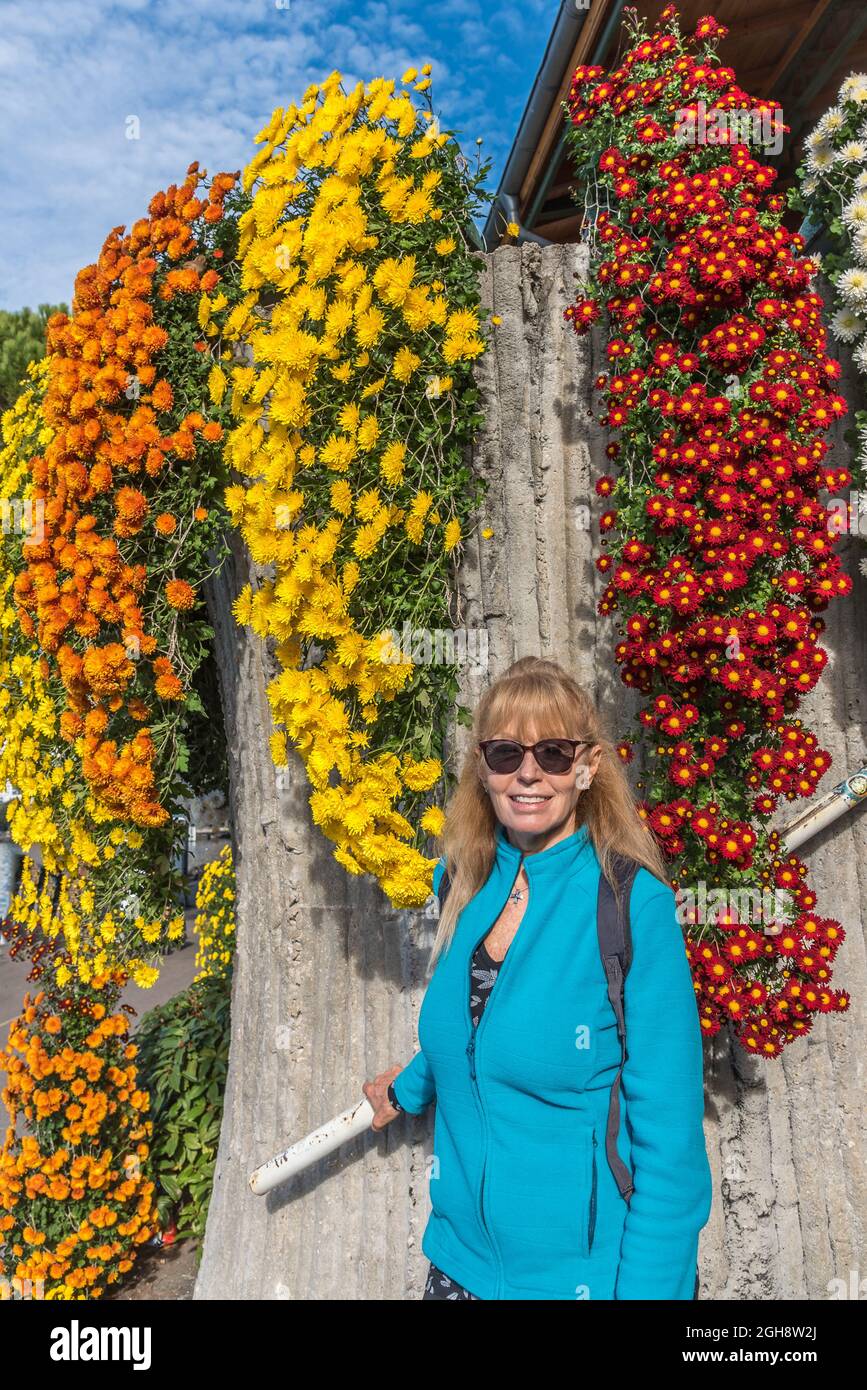 Vertikales Porträt einer erwachsenen blonden kaukasischen Frau, die vor einer Wand voller farbenprächtiger Blumen steht, sich an einem Geländer festhält und sich vor Th Stockfoto