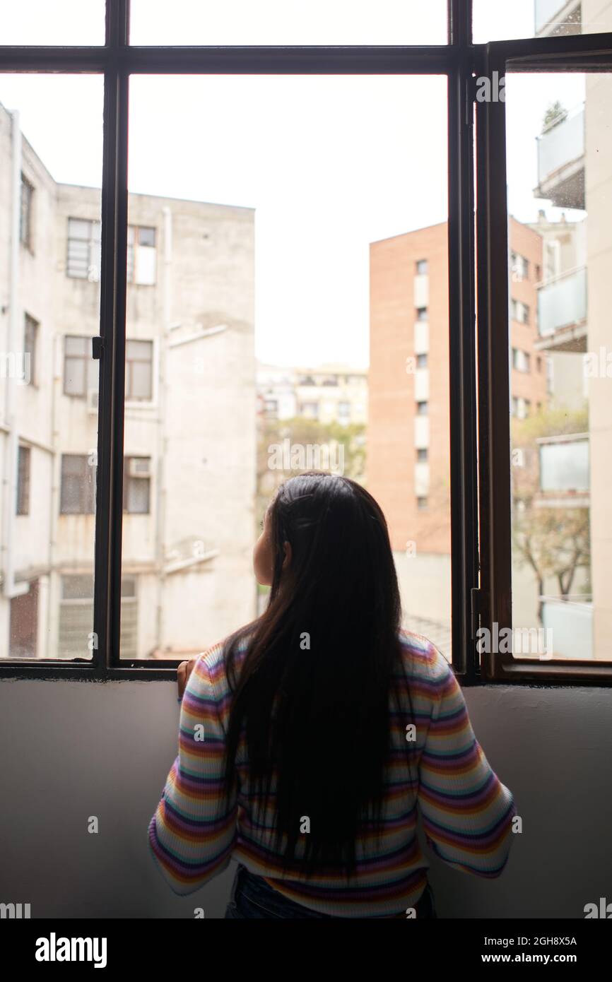 Vertikales Foto. Rückansicht afrikanisches Mädchen, das sich aus dem Fenster lehnte und nach draußen schaute, wo sich große Gebäude befinden. Stockfoto