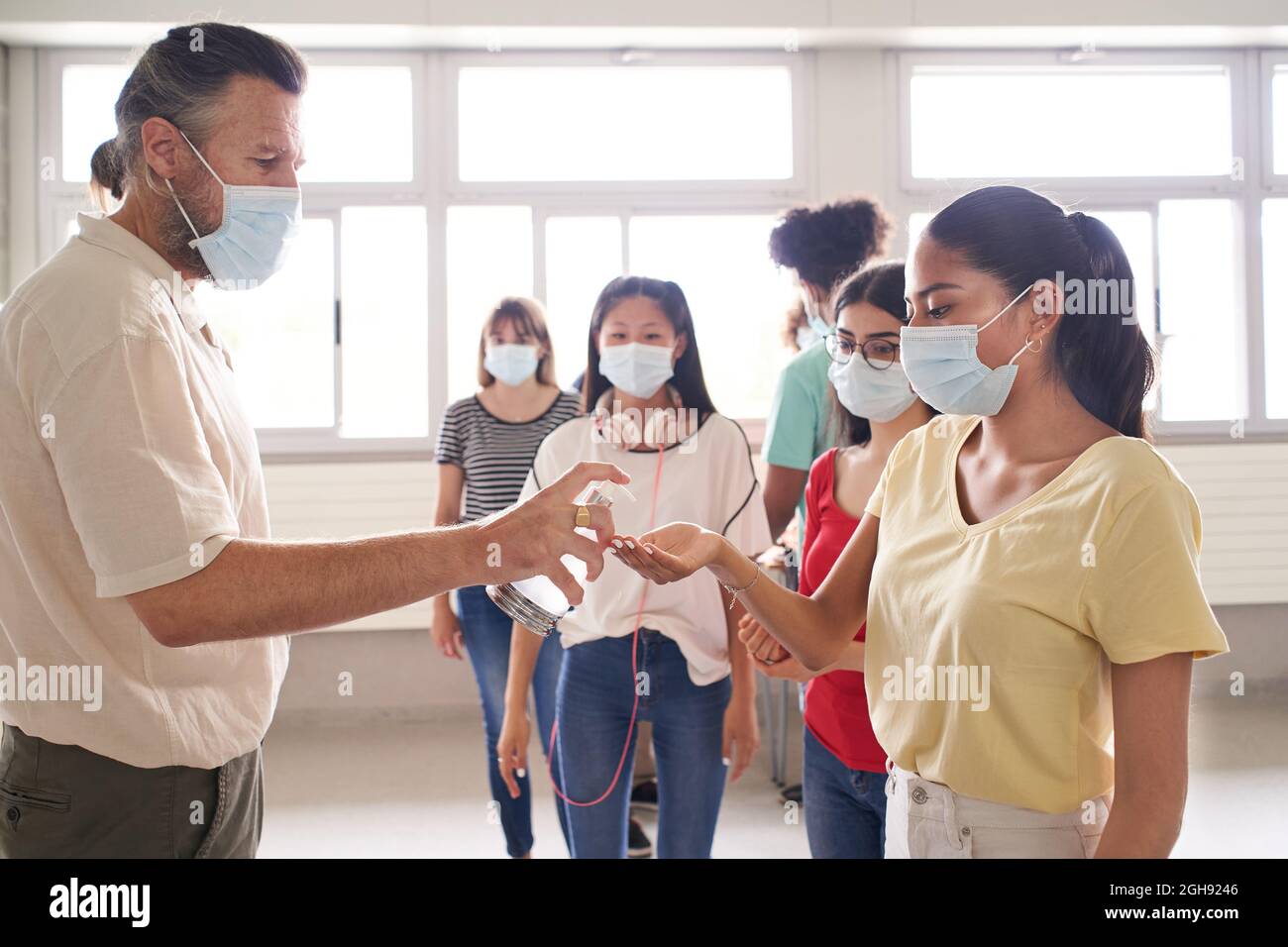Eine Gruppe von Schülern in Gesichtsmasken wartet in der Schlange, um in den Klassenzimmer zu kommen. Der Lehrer wendet hydroalkoholisches Gel für die Virenschutzmaßnahmen an. Zurück zur Schule. Stockfoto