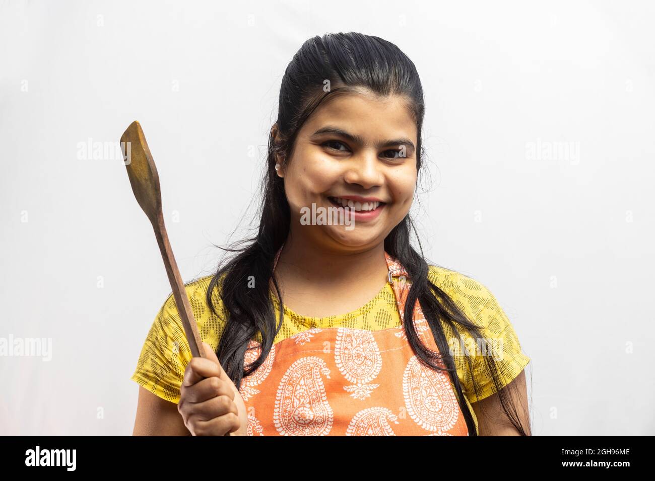 Eine hübsche indische Hausfrau in Kochschürze mit Holzspatel in der Hand lächelt auf weißem Hintergrund Stockfoto