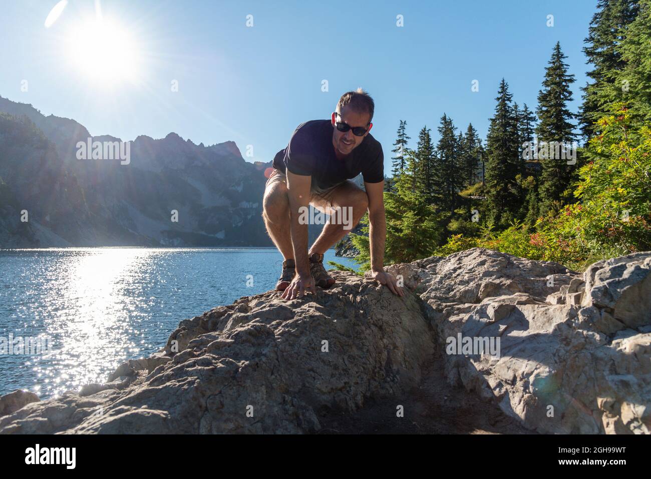 Lächelnder männlicher Wanderer, der über Felsen neben dem Bergsee klettert, spättägige Sonne. Stockfoto