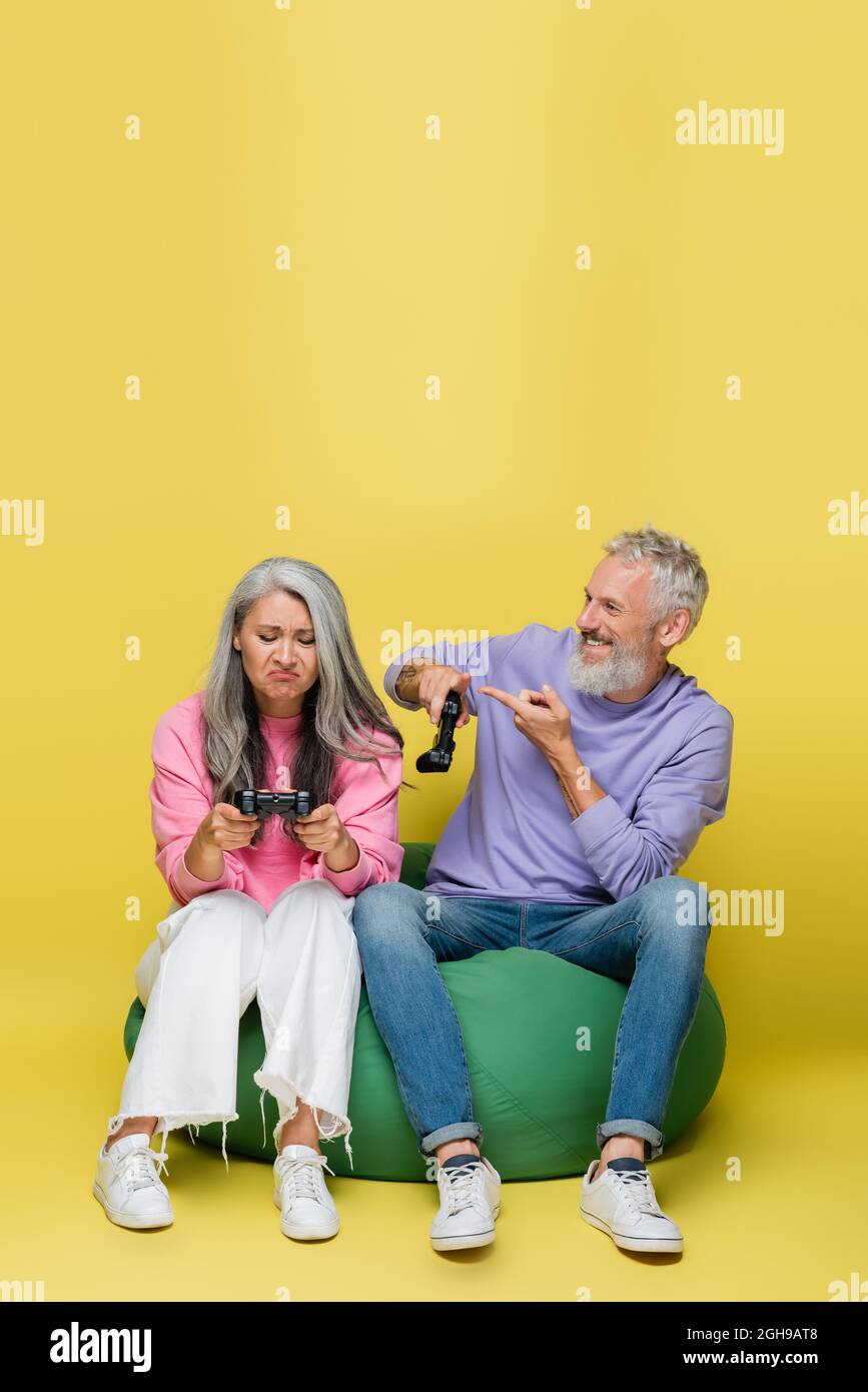 KIEW, UKRAINE - 10. AUGUST 2021: Glücklicher Mann mittleren Alters, der auf die traurige asiatische Frau zeigt, die ein Videospiel auf Gelb verliert Stockfoto