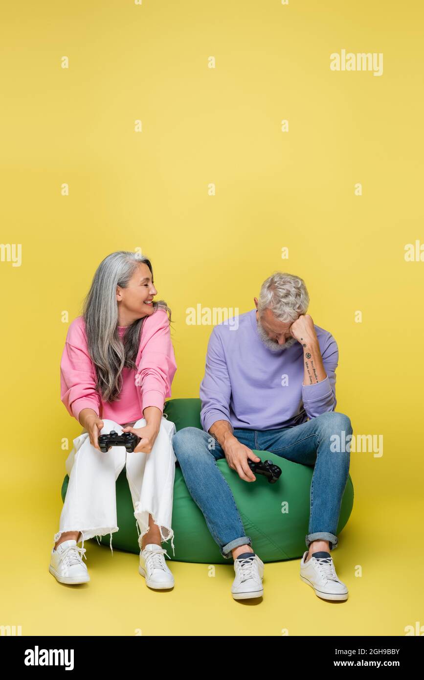 KIEW, UKRAINE - 10. AUGUST 2021: Glückliche asiatische Frau mittleren Alters mit Joystick in der Nähe traurigen Ehemann verlieren Videospiel auf gelb Stockfoto