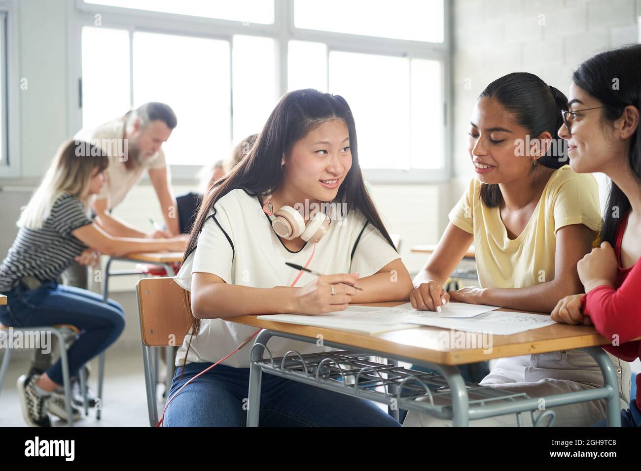 Lächelnde Studenten arbeiten im Unterricht als Team zusammen. Im Hintergrund hilft der Lehrer anderen Klassenkameraden. Stockfoto