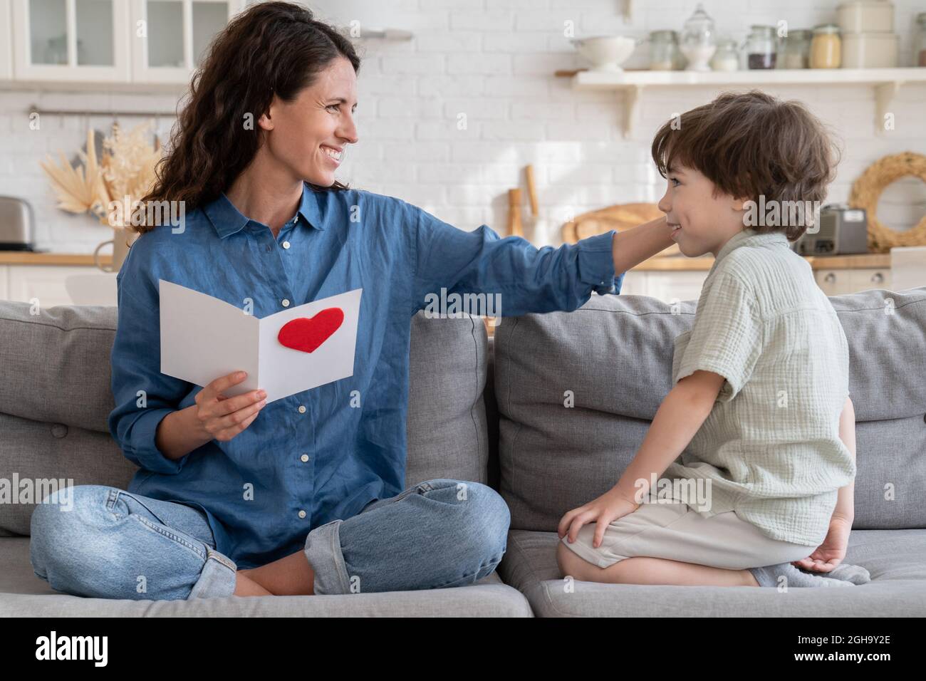 Dankbare Mutter berühren niedlichen Kind Sohn Haare halten Postkarte mit Grüßen am Muttertag sitzen auf der Couch zu Hause Stockfoto