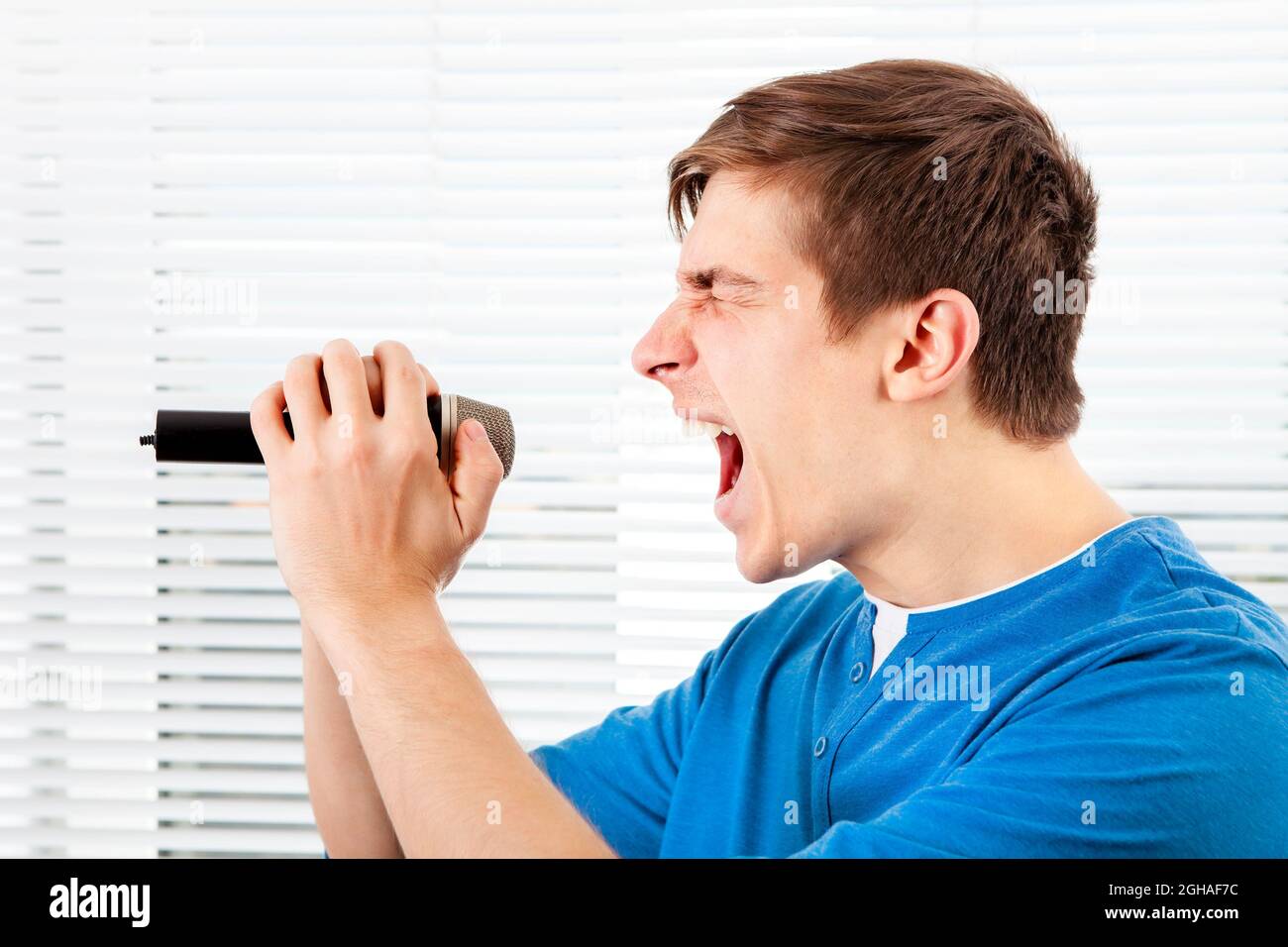 Junger Mann mit Mikrofon singt ein Lied auf dem Jalousie Hintergrund Stockfoto
