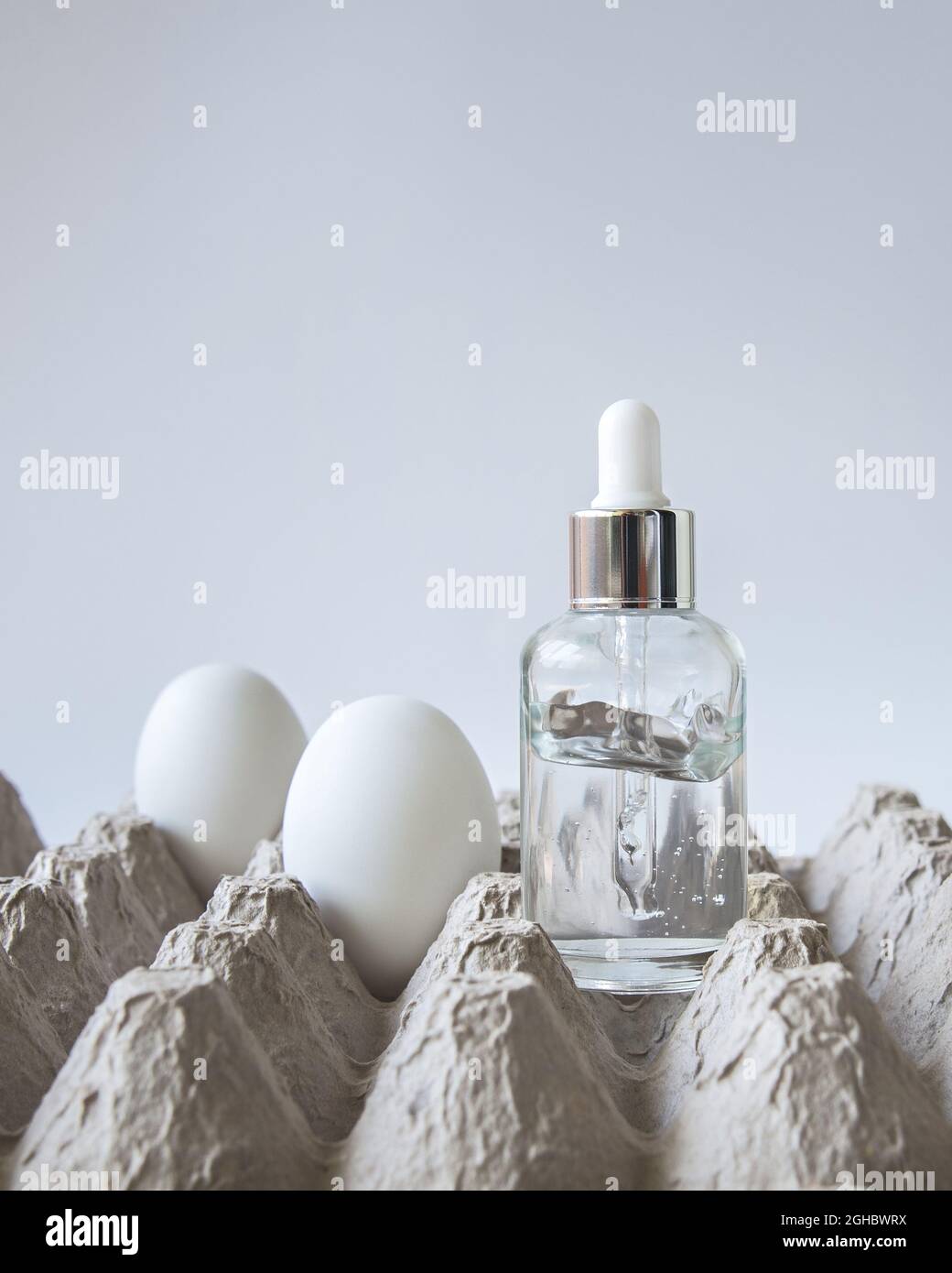 Auf einem Eierkarton steht eine Glasflasche mit Eiergesicht-Serum, in der Nähe befinden sich mehrere Eier auf dem weißen Hintergrund. Trendige natürliche Straffung. Stockfoto