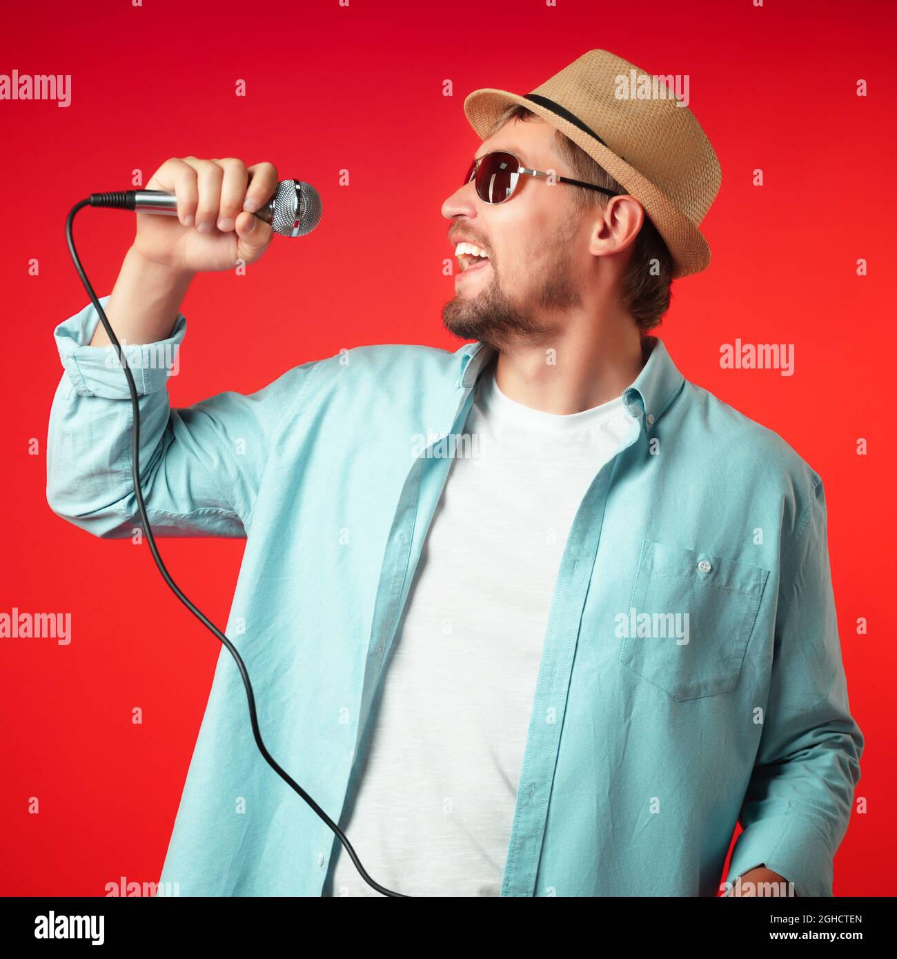 Ein Porträt eines Mannes mittleren Alters in einem Hut hält ein Mikrofon in der Hand und singt emotional ein Lied. Sänger in legerer Kleidung auf rotem Hintergrund. Stockfoto
