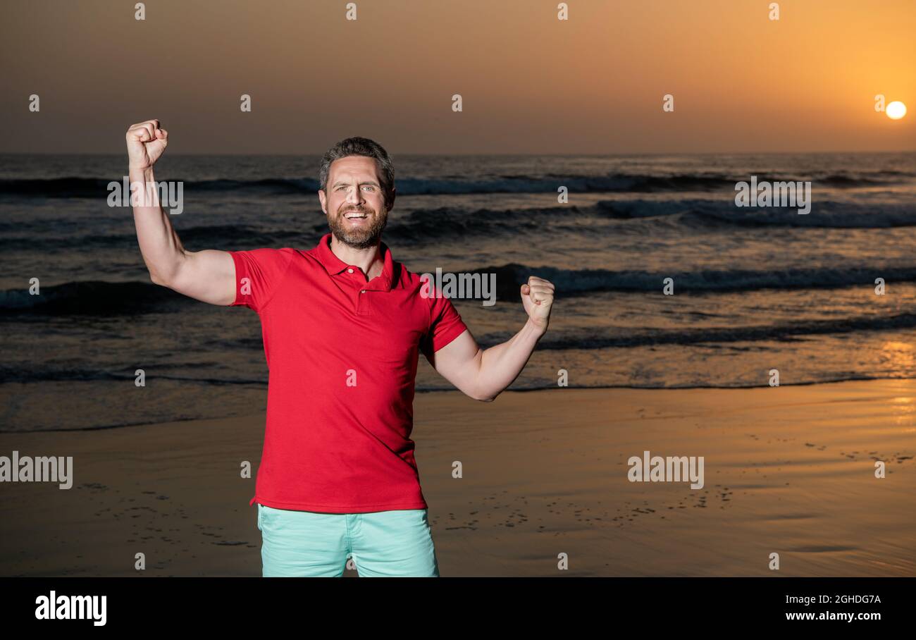 Erfolg feiern. Raum kopieren. Glücklicher Mann mit Bart im Sonnenuntergang über Meer. Erfolgreicher Kerl Stockfoto