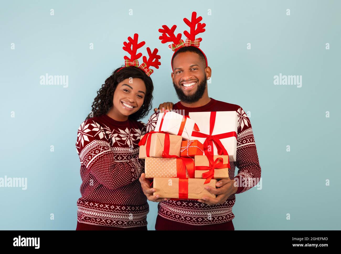 Glückliches schwarzes Paar mit Wollpullover und Hirschhörnern, Mann mit Haufen Weihnachtsgeschenke auf blauem Hintergrund Stockfoto