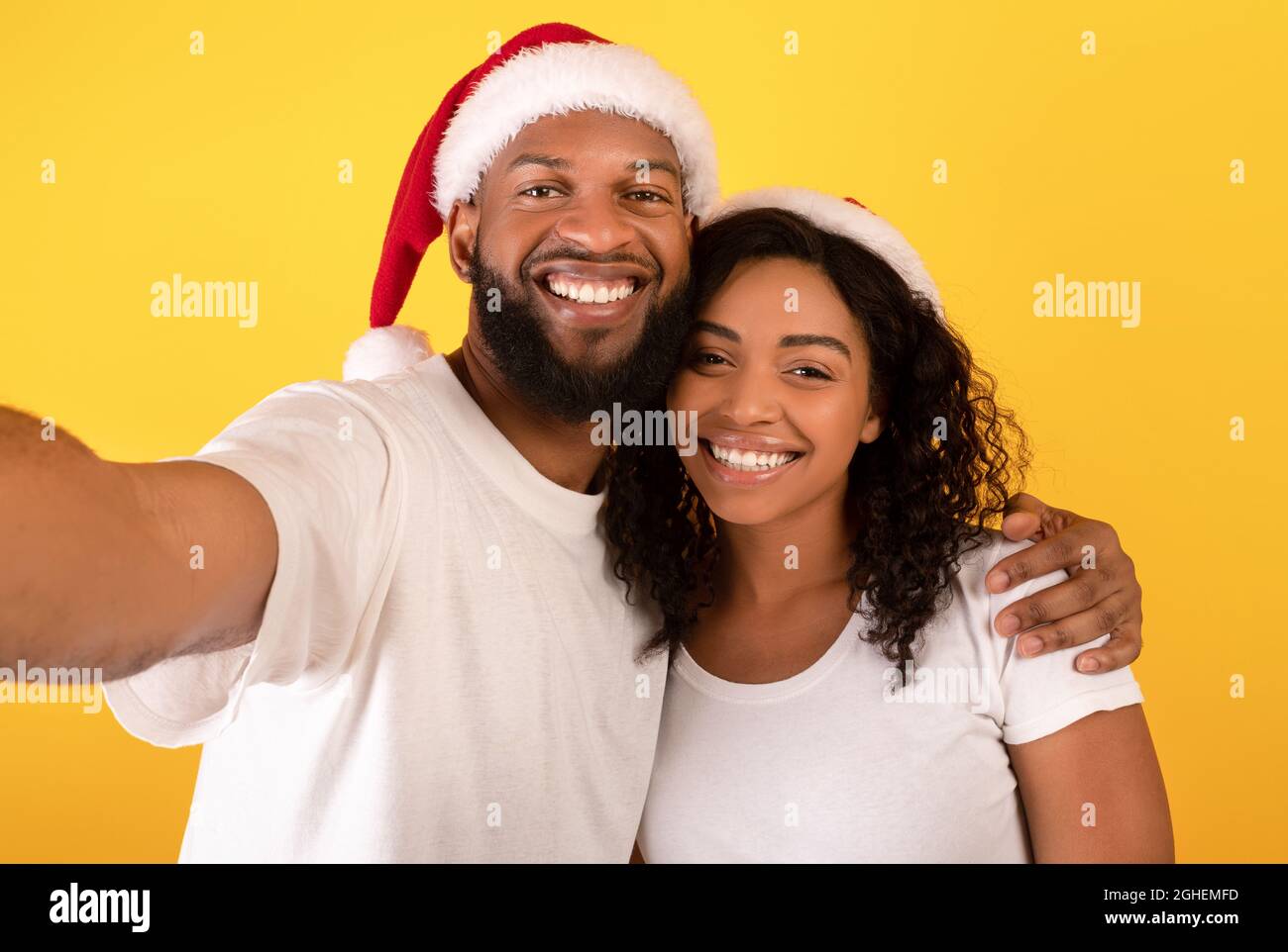 Liebevolles afroamerikanisches Paar in Weihnachtsmützen lächelnd und Selfie auf gelbem Studiohintergrund, freier Raum Stockfoto
