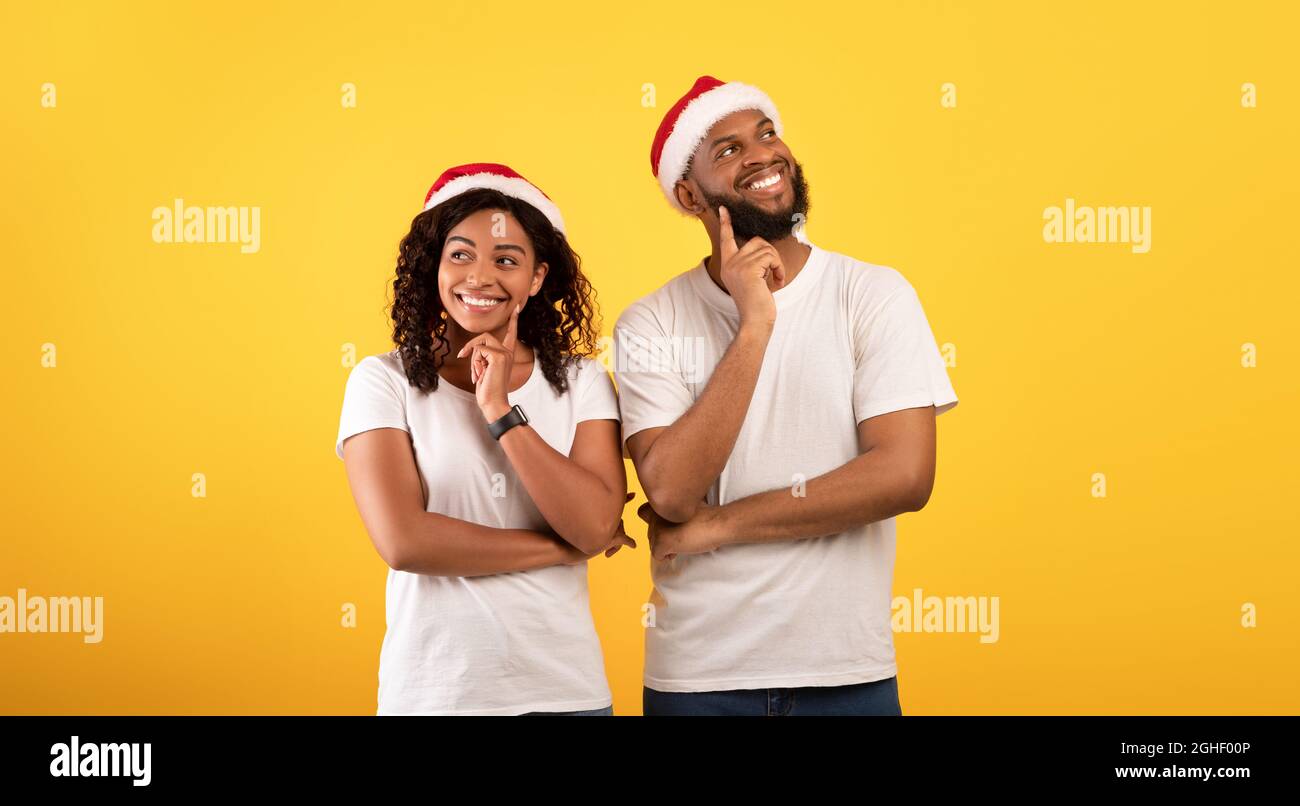 Verträumte afroamerikanische Ehepartner in Santa-Hüten, die die Finger auf die Chins halten und über den gelben Studiohintergrund nachdenken Stockfoto