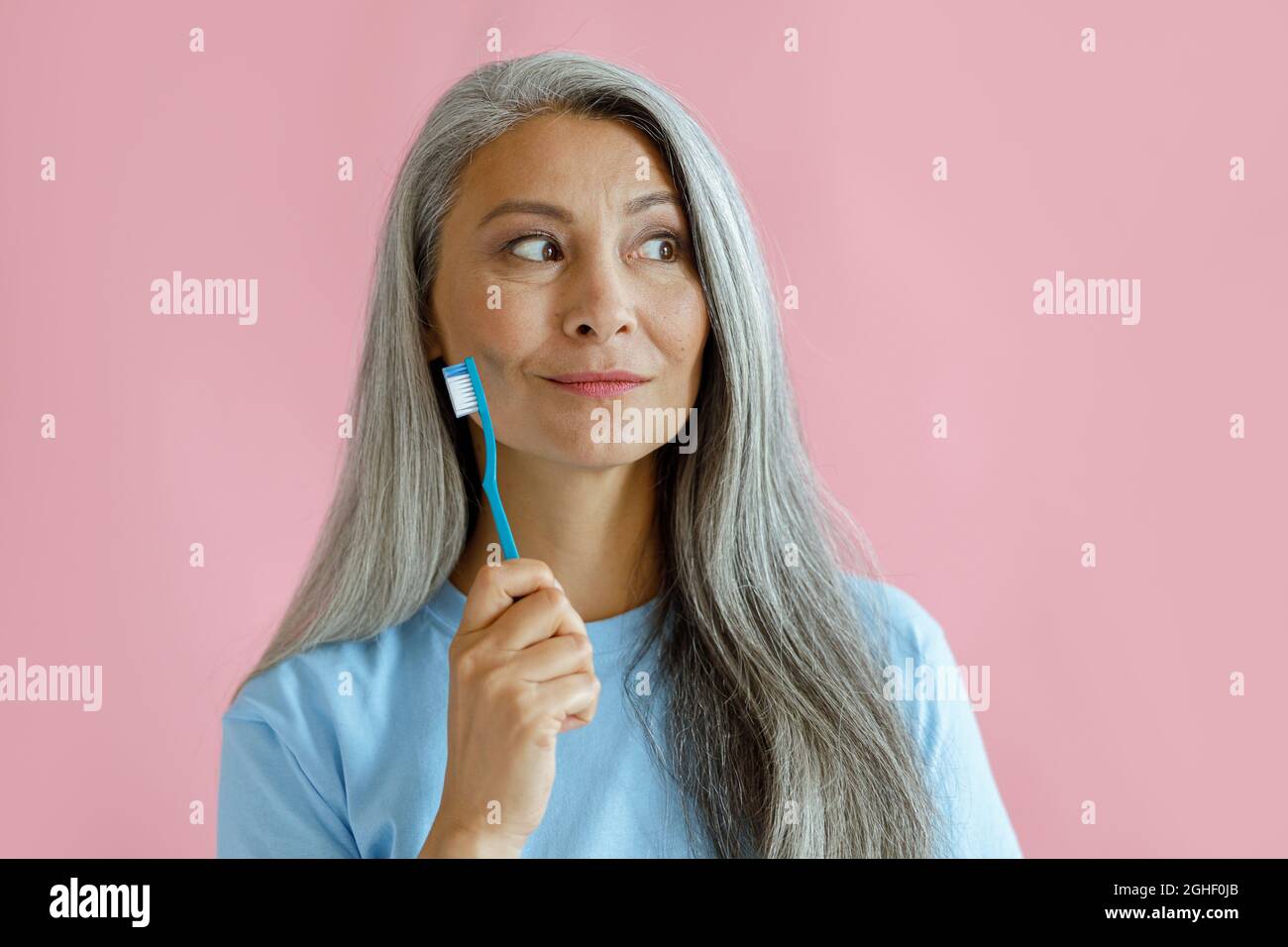 Positive nachdenkliche asiatische Frau mittleren Alters hält Zahnbürste im Studio Stockfoto