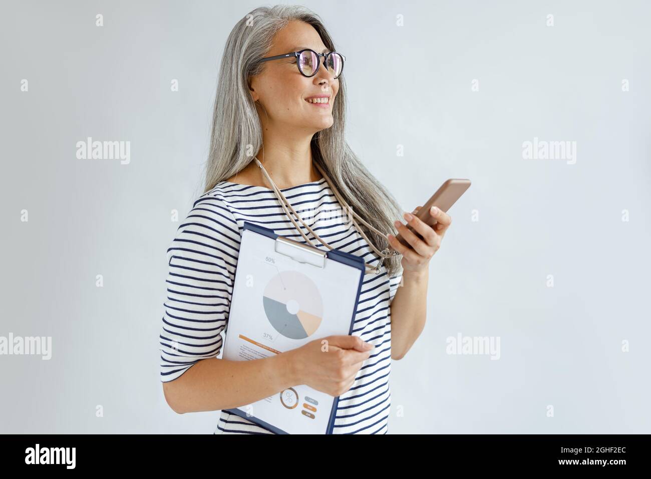 Reife asiatische Frau mit lockeren Haaren hält Diagramme und Handy auf hellgrauem Hintergrund Stockfoto