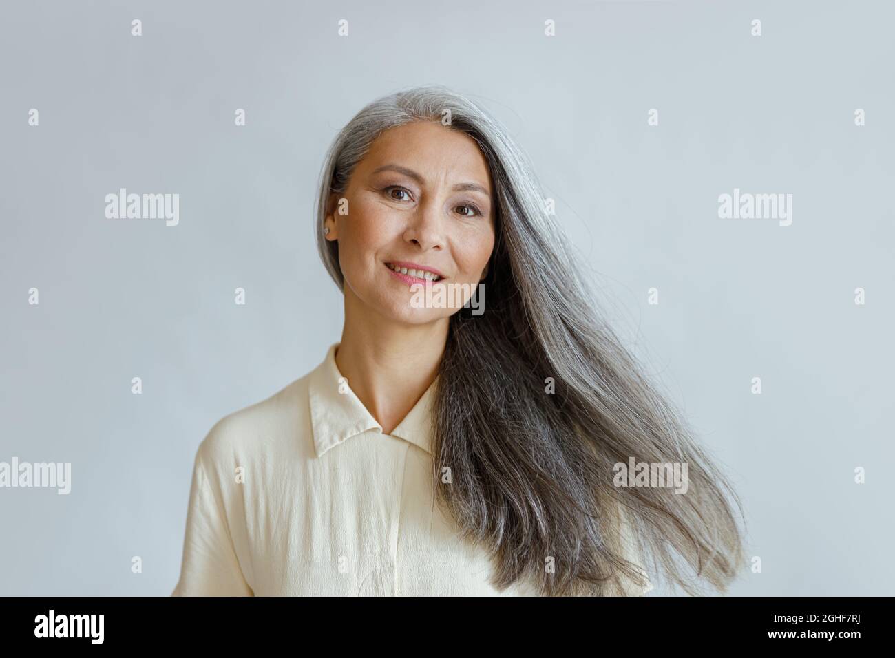 Lächelnde asiatische Frau mittleren Alters mit fließenden grauen Haaren steht auf hellem Hintergrund Stockfoto