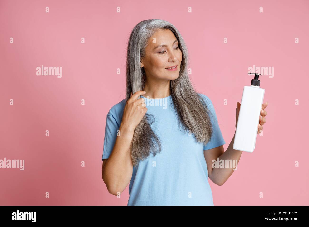 Schöne reife asiatische Frau mit silbernen Haaren hält leere Spenderflasche auf rosa Hintergrund Stockfoto