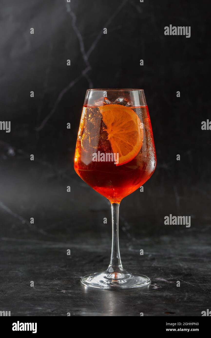 Ein Glas eisgekühlter Aperol Spritz-Cocktail, serviert in einem Weinglas, verziert mit Scheiben aus Orange und Basilikum, auf einem schwarzen Glas platziert Stockfoto