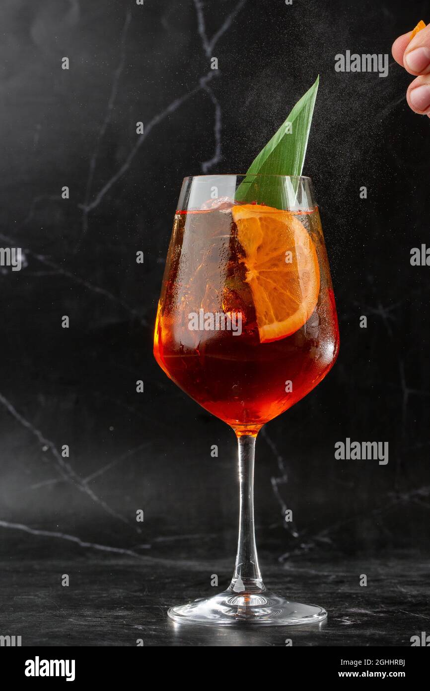 Ein Glas eisgekühlter Aperol Spritz-Cocktail, serviert in einem Weinglas, verziert mit Scheiben aus Orange und Basilikum, auf einem schwarzen Glas platziert Stockfoto